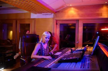 A young young engineer is sitting in a recording studio and using mixing console panel.
