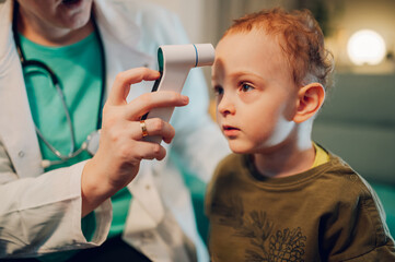 A boy is sitting at home while doctor is using thermometer on his forehead.