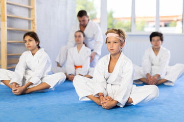 Fototapeta na wymiar Children in white kimono sits in a butterfly pose and practices stretching in sport gym