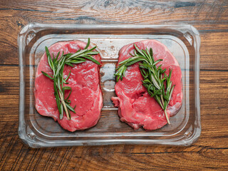 Juicy uncooked lamb leg steak with green fresh rosemary on plastic tray on wooden table. Uncooked high quality meat product with rich taste and vivid color.