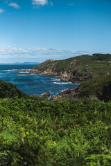 Captivating Ons Island scene: lush greenery in the foreground, azure sea, rocky cliffs, Pontevedra estuary, and a summer sky with scattered clouds from the Buraco do Inferno path.