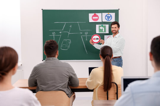 Teacher showing No Overtaking road sign to audience near chalkboard in driving school