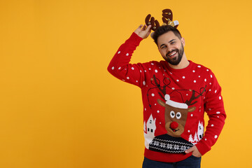 Happy young man in Christmas sweater and reindeer headband on orange background. Space for text - obrazy, fototapety, plakaty