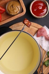 Fondue pot, forks with meat pieces and other products on black textured table, flat lay