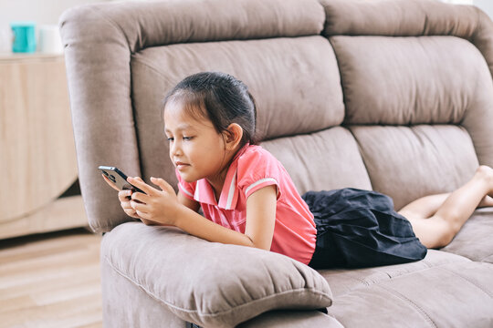 Smiling Little Girl Using Smartphone On Cozy Couch Having Fun Playing Game Or Watching Funny Video In Social Network