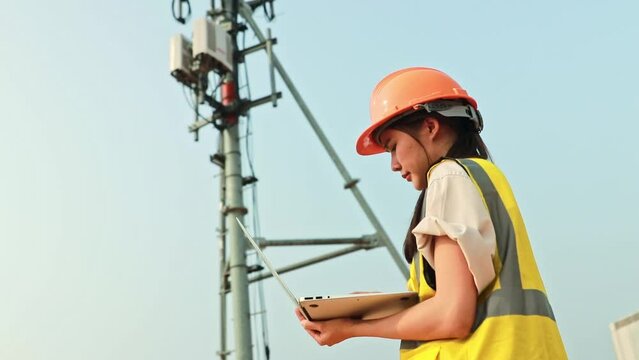 Asian female engineer wearing helmet performs field work telecommunications tower controlling mobile power installation using laptop maintain 5G network installed high rise building.