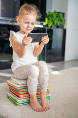 Cute Little girl sits on a stack of children's fairy-tale books and watches cartoons on her...