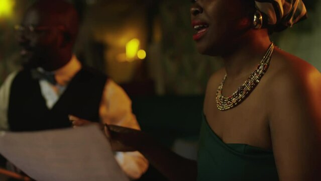 Waist up of artistic young African American woman wearing patterned headscarf and colorful beads necklace making beat with her fingers while singing son in chamber jazz club in evening