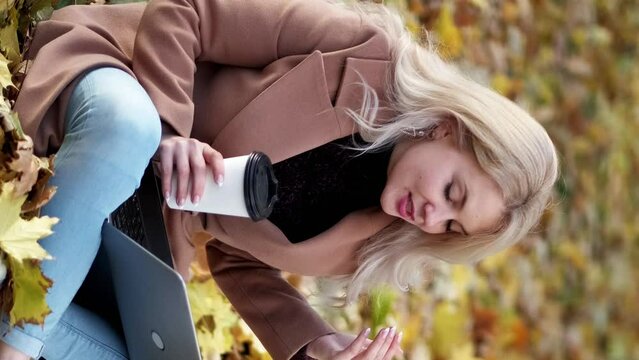 Vertical video. Online communication. Happy woman. Inspired autumn. Excited lady sitting on ground in park with cup coffee talking laptop showing yellow leaves.