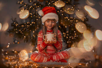 A portrait of a sweet beautiful smiling little 7-old girl wearing red pyjamas and Santas hat holding cup of kakao with candy in her hands at home near Christmas tree at Christmas eve. The concept of