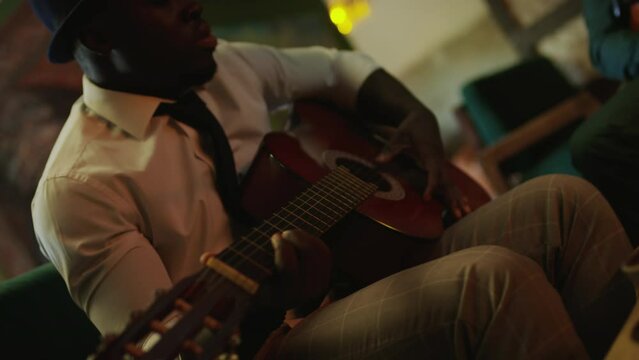 Young African American male jazz guitarist wearing white shirt, black tie and hat playing acoustic guitar while getting ready for evening performance in fancy jazz club