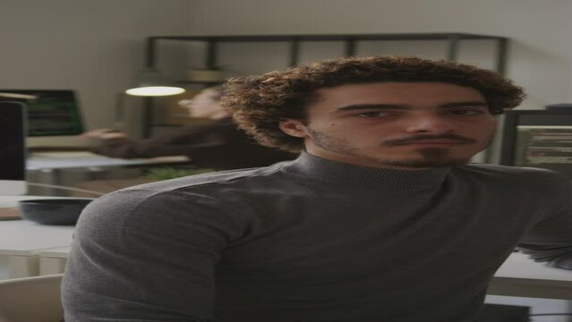 Vertical waist up portrait of curly haired Biracial male software engineer looking at camera seriously while sitting at workplace in contemporary office