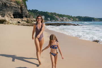 Beautiful little girl enjoying with her mother on the beach. They are running on the seashore and...