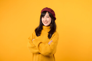 Satisfied young Asian woman in her 30s, confidently crossing her arms on her chest, dressed in a yellow sweater and red beret against a yellow background.