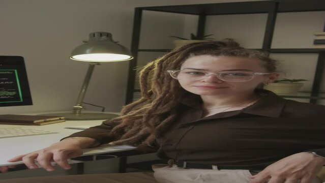 Vertical medium portrait of young Caucasian woman wearing eyeglasses and dreadlocks writing program code on wireless keyboard while working as software engineer in office
