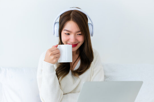 Morning Time in Bedroom Young Asian Woman Happy Drinking Milk and Working on Computer Laptop, Wearing headset, Sitting on Bed at House