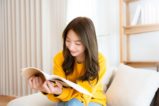 Young beautiful Asian woman reading a book on couch at home