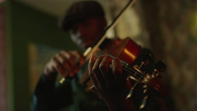 Defocused young Black man wearing dark green suit and black beret playing jazz violin in vintage style fancy music club with dimmed light