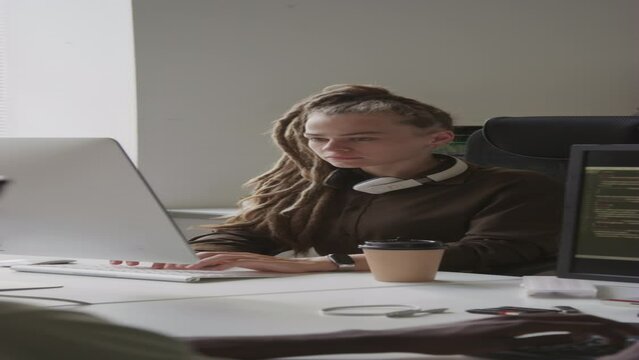 Vertical waist up shot of young Caucasian female software engineer with dreadlocks typing on wireless keyboard while working on desktop computer in office with Black male colleague