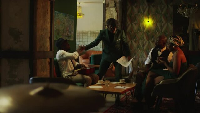 Full length shot of handsome young Black male jazz musician wearing dark green suit and beret arriving to rehearsal in fancy jazz club and greeting band members with handshake