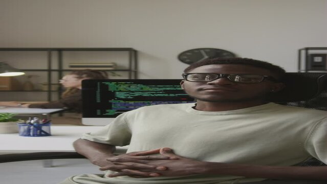 Vertical medium portrait of young Black male programmer sitting with his hands locked on stomach in chair at workplace in modern office and looking at camera