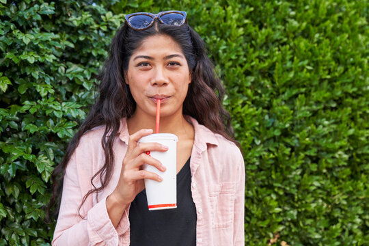 Woman drinks fast food soda with straw in front of hedge takes sip