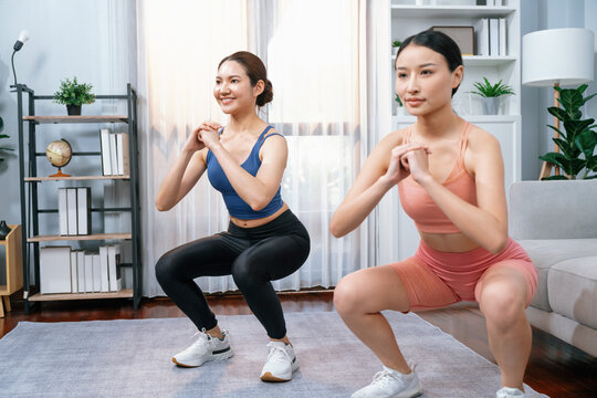 Vigorous energetic woman with trainer or workout buddy doing exercise at home. Young athletic asian woman strength and endurance training session as home workout routine with squat.