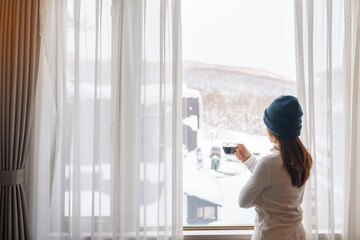 young woman in sweater with cup of coffee looking through the window in winter season, happy female enjoying snowfall outdoor view at apartment or home in the morning. Waking and Relaxing concept - Powered by Adobe