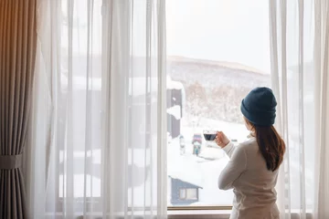 Foto op Aluminium young woman in sweater with cup of coffee looking through the window in winter season, happy female enjoying snowfall outdoor view at apartment or home in the morning. Waking and Relaxing concept © Jo Panuwat D