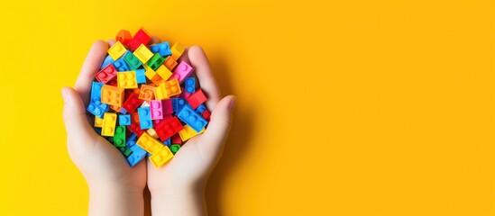 Hands holding colorful toy plastic bricks, blocks for building toys on yellow background - obrazy, fototapety, plakaty