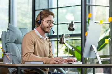 Handsome male programmer sits in a headset in the office at the table in profile, works at the computer, smiles and looks at the screen.