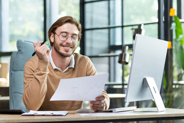 Satisfied young guy doing paperwork in the office, sitting at a desk in front of a computer, joyfully going through business papers, smiling