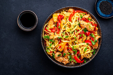 Stir fry noodles with chicken slices, pineapple, red paprika, chives, soy sauce and sesame seeds in ceramic bowl. Asian cuisine dish. Black table background, top view