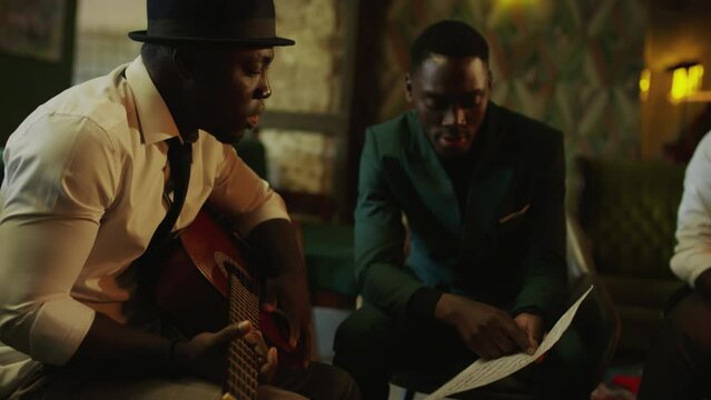 Medium shot of African American jazz quartet looking at music note sheet and discussing new song during rehearsal in fancy jazz club in evening