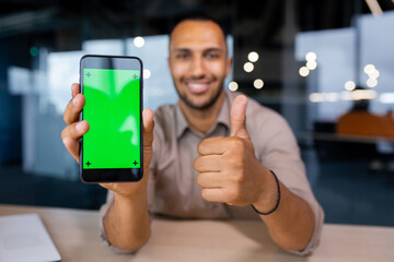 Happy man sitting in office holding phone with green screen and showing thumb up, phone in foreground.