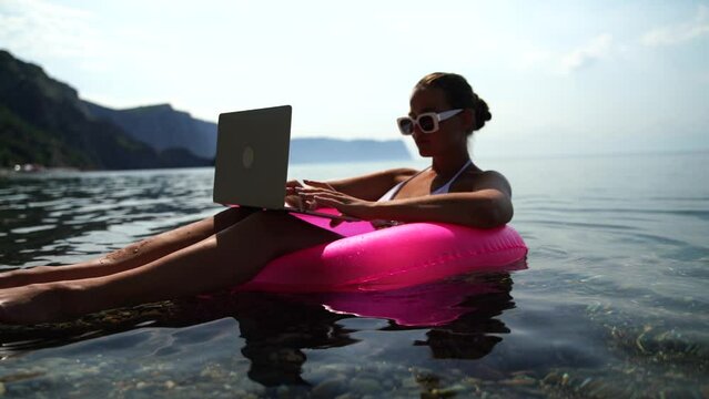Woman works on laptop in sea. Freelancer, blond woman in sunglases floating on an inflatable big pink donut with a laptop in the sea at sunset. People summer vacation rest lifestyle concept.