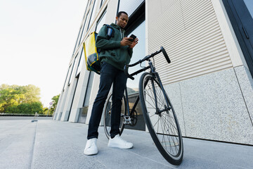 A dark-skinned deliveryman with a backpack stands next to a bicycle against the background of an office building. The courier holds a smartphone in his hands.