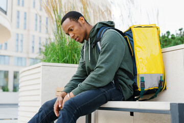 A tired and tortured courier is sitting on a bench after a work shift. African American delivery...