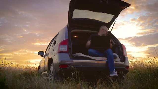 Tired adult man hate workforce life hybrid digital work anywhere turn off close logout laptop after end hard job done, sitting in car trunk while traveling, general plan in field at sunset. Busy