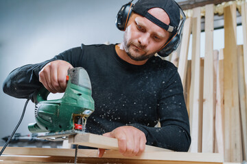 A carpenter man in dark clothes cutting a board with an electric jigsaw. Manufacture of furniture...