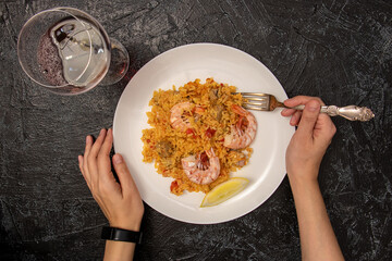 top view of female hands with fork, white plate with paella and shrimp and glass of red wine on black textured background. dinner alone.