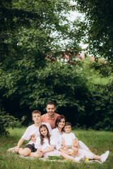 Naklejka premium Big family sitting on the picnic blanket in city park during weekend Sunday sunny day. They are smiling, laughing and eating boiled corn and watermelon.