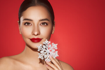 Beautiful young woman with red lipstick, shiny clean skin and face, showing glowing snowflake, near face and smiling, isolated on a red background. Christmas holidays concept