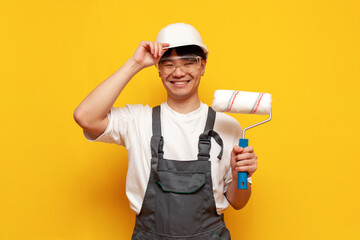 Asian male painter in helmet and overalls holds roller and smiles on yellow isolated background,...