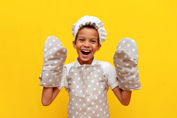 boy african american baker in uniform and chef's hat shows thermal gloves for baking on yellow isolated background - obrazy, fototapety, plakaty