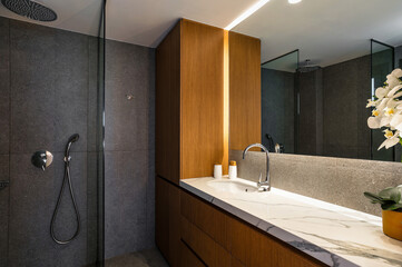 Modern Luxury Bathroom: Stylish Grey Walls, Rain Shower, Brown Wooden Cabinet, White Sink and Expansive Wall Mirror. Ideal for use in interior design concepts, home deco publications, and presentation