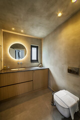 Modern bathroom interior design featuring brown and yellow color walls, a white toilet bowl, a sink on a wooden stand, and a round mirror on the wall with backlighting