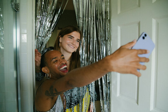 Young non-binary person and woman taking selfie with silver tinsel curtain in the bathroom