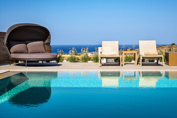Summer sunny day: Double sun sofa with shade and two sunbeds next to the blue water pool, with a blurred panoramic sea view in the background