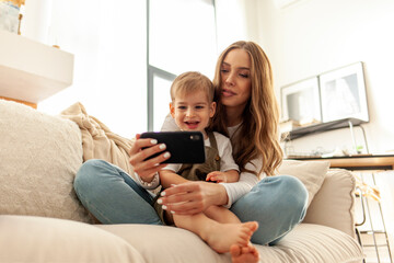 young mother uses smartphone with her little son at home on the sofa, 2-year-old boy looks at mobile phone